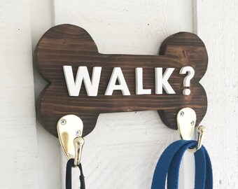 Op de afbeelding: Een houten bord in de vorm van een hondenbot met de tekst "WALK?" in witte letters. Aan het bord zijn twee haken bevestigd, waaraan een zwarte riem aan de ene en een blauwe riem aan de andere hangt.