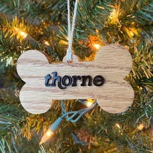 Op de afbeelding: Een houten hondenbot ornament met de naam "Thorne" erop geschreven in zwarte letters. Het ornament hangt aan een kerstboom met witte lampjes.