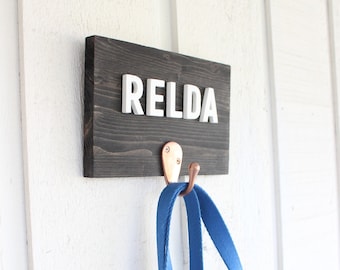 Op de afbeelding: Een donkerbruin houten bord met de naam "RELDA" in witte letters. Een koperen haak is bevestigd aan het bord met een blauwe riem die eraan hangt.