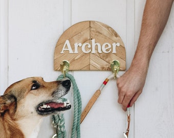 May include: A wooden dog leash holder with the name "Archer" in white lettering. The holder has two brass hooks and is mounted on a white door. A dog and a leash are visible.