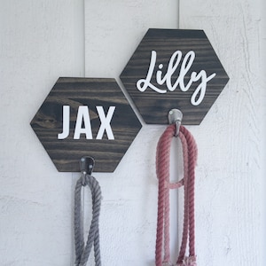May include: Two wooden hexagon signs with the names "Jax" and "Lilly" written in white lettering. Each sign has a metal hook attached to the bottom, with a rope leash hanging from each hook.
