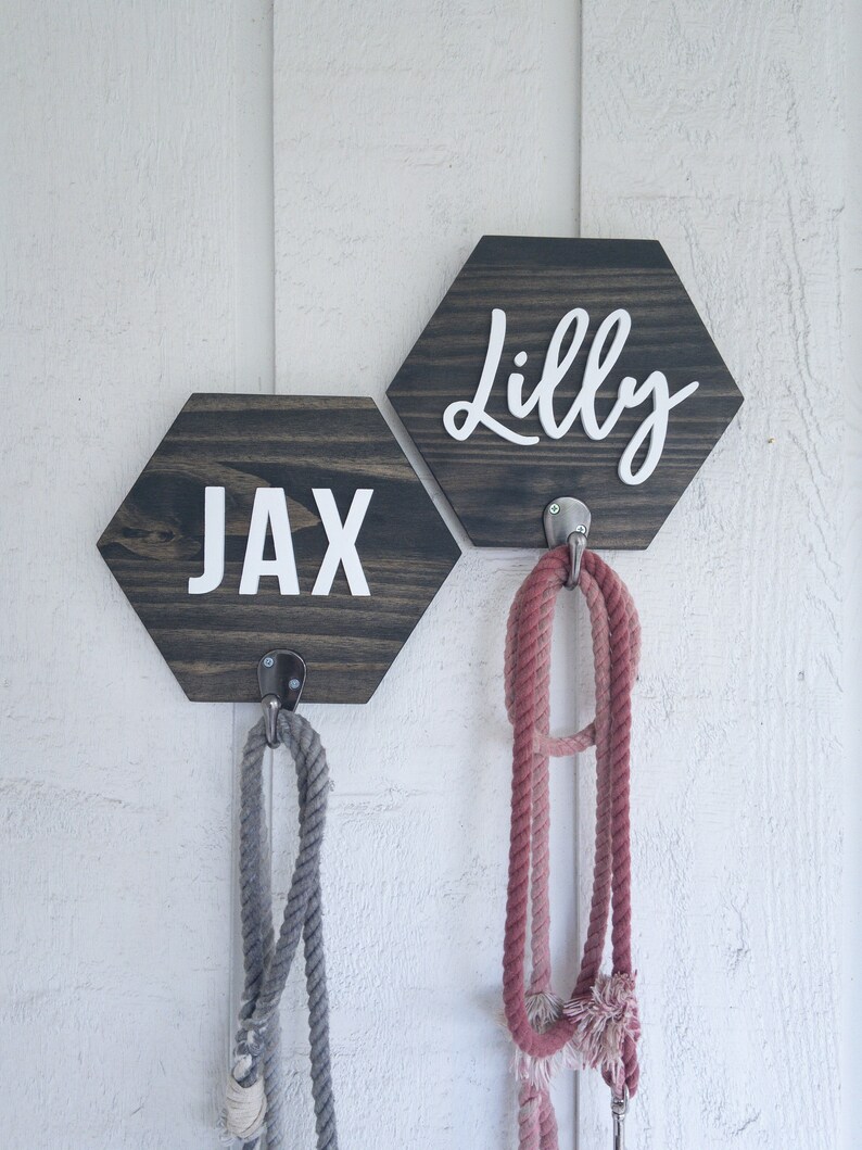 May include: Two wooden hexagon signs with the names "Jax" and "Lilly" written in white lettering. Each sign has a metal hook attached to the bottom, with a rope leash hanging from each hook.