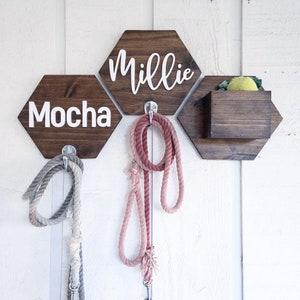 May include: Three wooden hexagonal wall hooks with the names "Millie", "Mocha", and a small wooden box with a yellow toy inside. Each hook has a silver metal hook for hanging leashes. Two leashes are hanging from the hooks, one pink and one gray.
