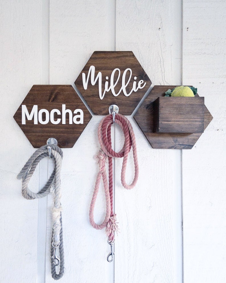 May include: Three wooden hexagonal wall hooks with the names "Millie", "Mocha", and a small wooden box with a yellow toy inside. Each hook has a silver metal hook for hanging leashes. Two leashes are hanging from the hooks, one pink and one gray.