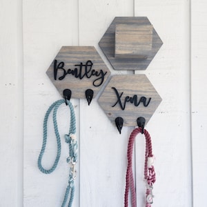 May include: Two wooden hexagon-shaped dog leash holders with hooks. The holder on the left says "Bentley" and the holder on the right says "Xena". A blue leash is hanging on the left hook and a pink leash is hanging on the right hook.