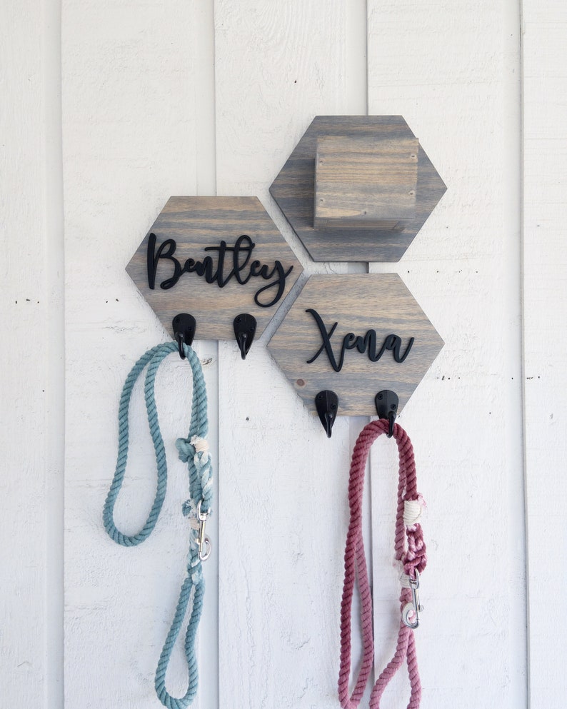 May include: Two wooden hexagon-shaped dog leash holders with hooks. The holder on the left says "Bentley" and the holder on the right says "Xena". A blue leash is hanging on the left hook and a pink leash is hanging on the right hook.
