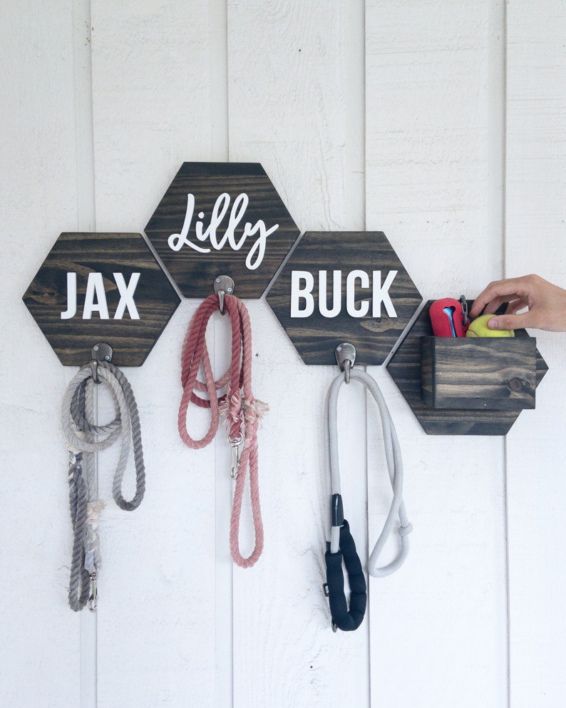 May include: A wooden dog leash holder with three hexagon-shaped hooks and a small storage compartment. The hooks are labeled "Jax", "Lilly", and "Buck".