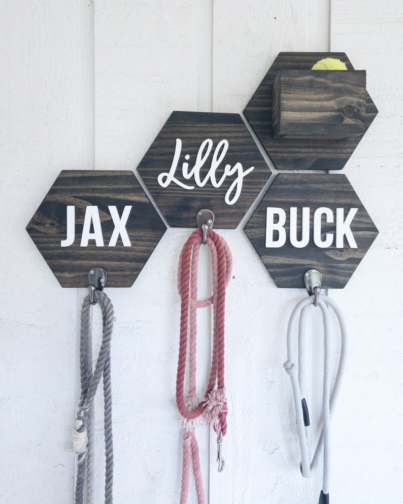 May include: A wooden wall organizer with three hexagonal shelves, each with a dog's name on it: Jax, Lilly, and Buck. Each shelf has a hook for a leash, and there is a small wooden box for storing dog toys.