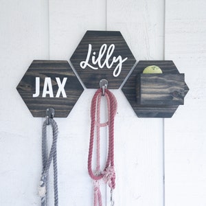 May include: A wooden dog leash holder with two hexagon-shaped signs that say "Jax" and "Lilly" and a small wooden box for storing dog treats. The holder has two hooks for hanging leashes.