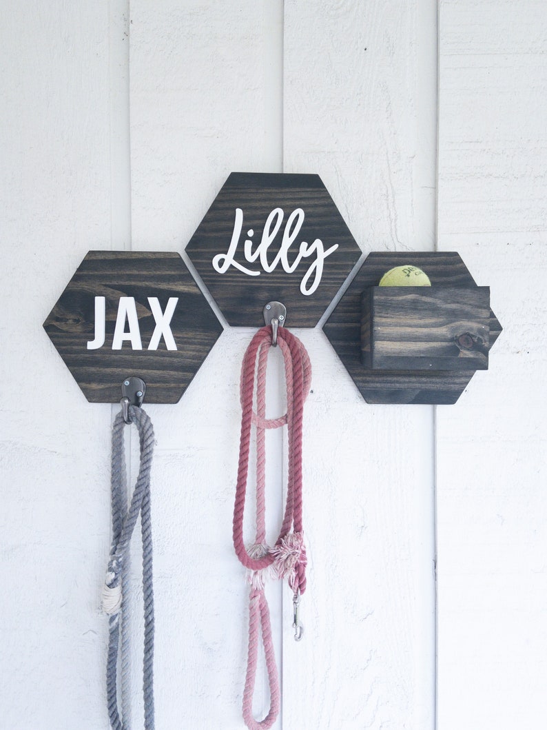 May include: A wooden dog leash holder with two hexagon-shaped signs that say "Jax" and "Lilly" and a small wooden box for storing dog treats. The holder has two hooks for hanging leashes.