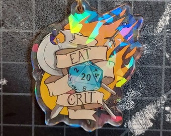 Könnte beinhalten: Ein holografischer Schlüsselanhänger mit einem blauen 20-seitigen Würfel mit den Worten "Eat Crit" auf einem Banner. Der Würfel ist von zwei Schwertern und einem Flammenmuster umgeben.