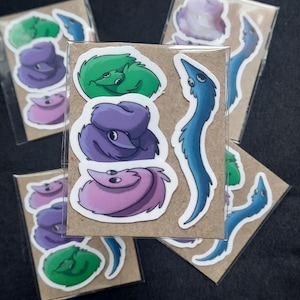 May include: A set of five worm on a string stickers. The stickers are on a brown background and feature different colored worms with googly eyes. The worms are green, purple, blue, and pink.