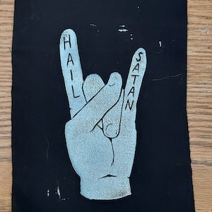 K&ouml;nnte beinhalten: Ein schwarzer Stoff-Patch mit einer wei&szlig;en Hand, die die Rock'n'Roll-Handgeste macht. Die Worte "HAIL SATAN" sind auf den Fingern geschrieben.