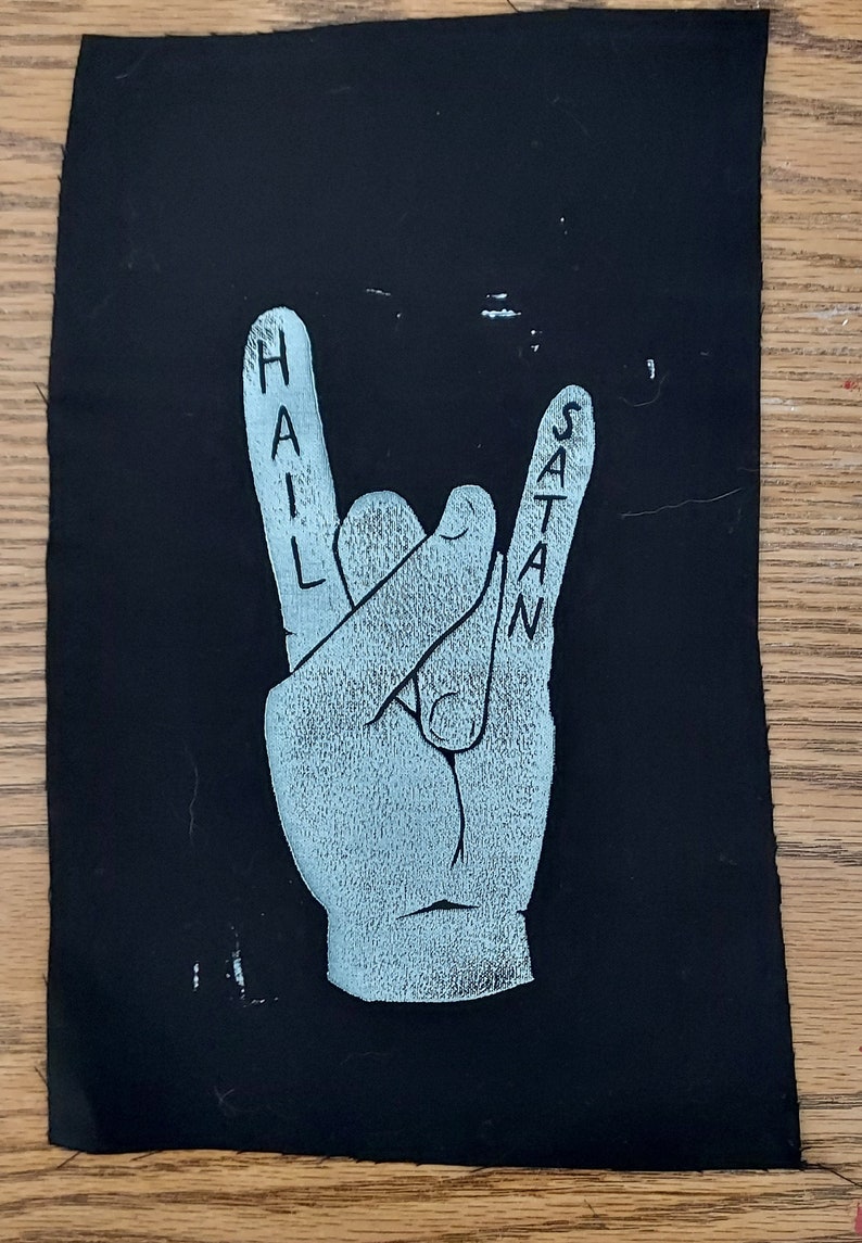 K&ouml;nnte beinhalten: Ein schwarzer Stoff-Patch mit einer wei&szlig;en Hand, die die Rock'n'Roll-Handgeste macht. Die Worte "HAIL SATAN" sind auf den Fingern geschrieben.
