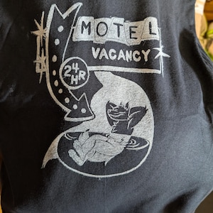 May include: Black t-shirt with a distressed white graphic. The graphic features a motel sign with the words "MOTEL VACANCY" and a 24-hour symbol. Below the sign is an illustration of a creature and a hand.