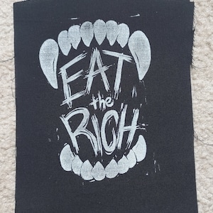 K&ouml;nnte beinhalten: Schwarzer Stoff-Patch mit wei&szlig;em Text, der "Eat the Rich" innerhalb einer wei&szlig;en Grafik eines Gebisses lautet.