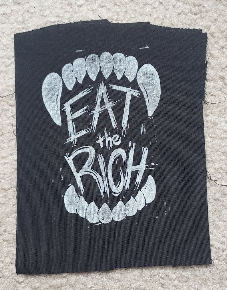 K&ouml;nnte beinhalten: Schwarzer Stoff-Patch mit wei&szlig;em Text, der "Eat the Rich" innerhalb einer wei&szlig;en Grafik eines Gebisses lautet.