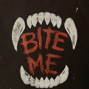 K&ouml;nnte beinhalten: Eine schwarze Stofftasche mit einem wei&szlig;en Umriss eines Gebisses und den Worten "BITE ME" in Rot.