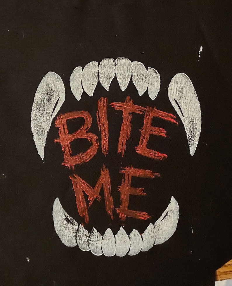 K&ouml;nnte beinhalten: Eine schwarze Stofftasche mit einem wei&szlig;en Umriss eines Gebisses und den Worten "BITE ME" in Rot.