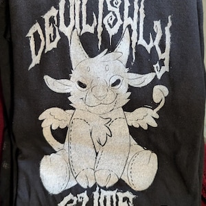 Puede incluir: Camiseta gris oscuro con un gráfico plateado de una criatura demoníaca de dibujos animados y las palabras "DEVILISHLY CUTE". El diseño presenta un diablillo sonriente con alas. Una etiqueta "M" es visible.