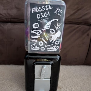May include: A black gumball machine with a clear top, featuring a "Fossil Dig!" sign with dinosaur fossil illustrations. The sign indicates "3 small patches in each capsule". The machine has a silver coin slot and a crank.