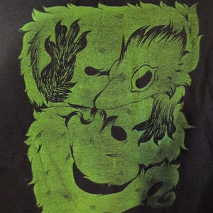 May include: Black t-shirt with a green graphic of a cartoonish creature. The design shows a smiling face, claws, and a furry outline. The artwork is printed on the front of the shirt.