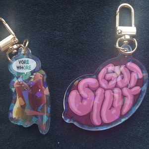 May include: Two iridescent keychains with gold metal rings. One keychain features a pink cartoon brain with the text "GO! SLUT" in pink. The other keychain features a cartoon red and yellow creature with the text "VORE WHORE" in a speech bubble.