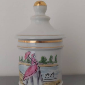 Può includere: Barattolo in porcellana bianca con coperchio e bordo dorati. Il barattolo presenta una scena dipinta a mano di una donna in un abito rosa che cammina con un ombrello. La scena è ambientata sullo sfondo di un lago e alberi. Il barattolo è etichettato "Au Sucre".