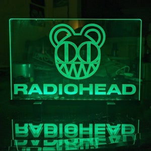 May include: A green neon sign with the Radiohead logo and the band's name in white letters. The logo is a stylized cartoon face with two large eyes and a jagged mouth.