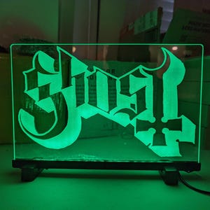 May include: A glowing green LED sign featuring the band name "Ghost" in a gothic font with a cross symbol. The sign is rectangular, mounted on a black base, and emits a bright green light.