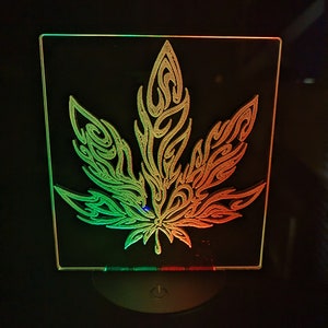Puede incluir: Una luz nocturna de acrílico transparente con un diseño estilizado de hoja de cannabis. La luz se ilumina en tonos de verde, amarillo y rojo.