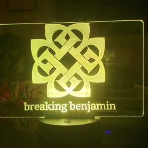 May include: A yellow illuminated acrylic sign with a stylized logo of a four-leaf clover and the text "breaking benjamin" below.