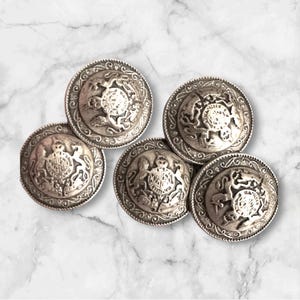 May include: Five antique silver-colored buttons with ornate detailing. Each button features a central emblem surrounded by a decorative border. The buttons are round and have a slightly aged appearance, suggesting vintage craftsmanship.