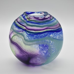 May include: A round glass vase with a swirling pattern of purple, blue, and green. The vase has a clear glass base and a white interior.