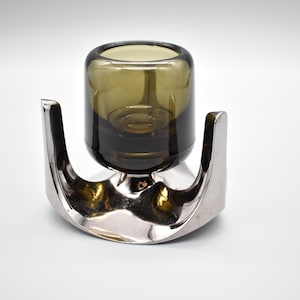 Peut inclure: Un bougeoir moderne avec une bougie votive en verre vert olive foncé, placée dans un support en métal argenté poli. La base métallique a une forme incurvée et organique. Conçu pour une seule bougie.