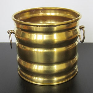 Könnte beinhalten: Ein goldfarbener Metall-Pflanztopf mit zwei Griffen. Der Topf hat ein geripptes Design und eine glänzende Oberfläche.