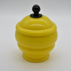 May include: A yellow glass jar with a black knob lid. The jar has a rounded shape with a series of horizontal ridges.