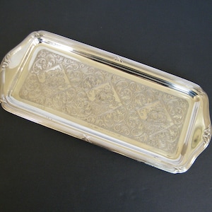 May include: A rectangular silver tray with detailed floral and scroll patterns. The tray features a raised, ribbed edge and decorative corners. The surface is highly reflective, and the design is elegant and classic. The tray is likely made of silver.