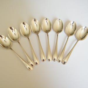 May include: A set of eight polished silver-toned metal spoons arranged in a fan-like formation. Each spoon features a smooth, rounded bowl and a slender, plain handle with a slightly tapered end, reflecting light across their metallic surfaces.