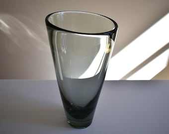 May include: A tall, gray glass vase with a unique, angled rim. The vase has a sleek, modern design, with a dark base that fades to a lighter gray towards the top. The interior is white, and the vase is empty.