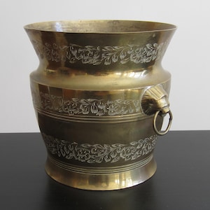 Peut inclure: Un pot en laiton avec un motif floral et des poignées en tête de lion. Le pot est de couleur or clair et a une finition brillante.
