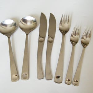 May include: A collection of stainless steel cutlery, comprising two spoons, two knives, and three forks. The utensils feature a minimalist design with a circular detail at the handle's base. The cutlery is displayed on a white surface.