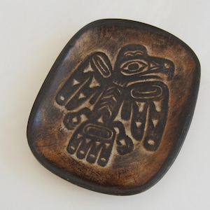 Puede incluir: Un plato de cerámica marrón con un diseño en relieve de un águila estilizada en negro. El águila es un motivo de arte tradicional de los pueblos indígenas de la costa noroeste.
