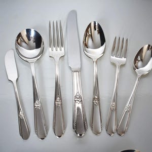 May include: A collection of polished silver cutlery, including spoons, forks, and knives, displayed on a light grey surface. The silverware features decorative detailing on the handles. The set includes a butter knife. Suitable for formal dining.