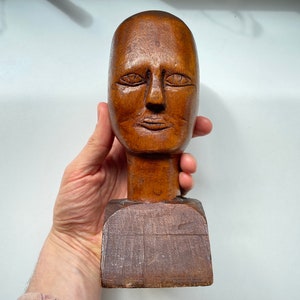 May include: A wooden sculpture of a human head with a simple, carved face. The head is mounted on a wooden base.