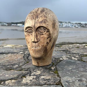 May include: A carved wooden head sculpture with a serene expression. The sculpture is light brown with visible wood grain and a vertical split down the center. It is placed on a stone surface with a cloudy sky in the background.