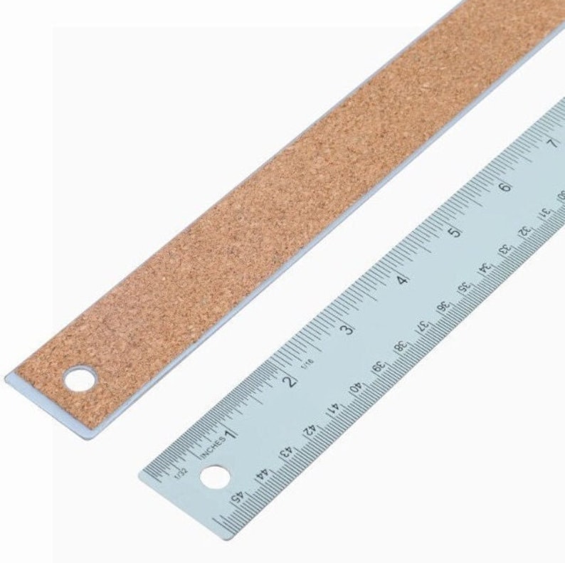 May include: A silver ruler with cork backing. The ruler is marked in inches and has a hole at the 1 inch mark and another hole at the 6 inch mark.