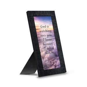 May include: Black framed picture with a quote that reads "God is watching over you. I know because I asked Him to." The picture features a cloudy sky with a sunset in the background.