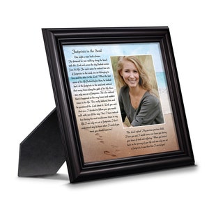 May include: A black framed picture with a white mat and a photo of a woman smiling. The picture has a quote about footprints in the sand and a depiction of footprints in the sand.