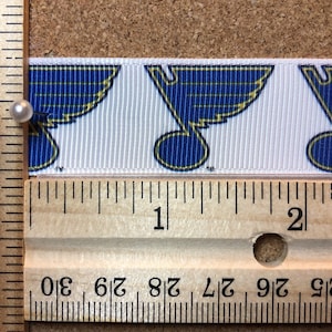 May include: A white ribbon with a repeating pattern of blue and gold hockey team logos. The ribbon is measured against a ruler, showing it is 1 inch wide.